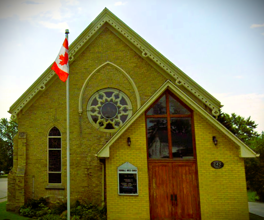 a yellow brick church with wooden doors a flagpole with a canadian flag in the foreground and in the background blue sky and trees