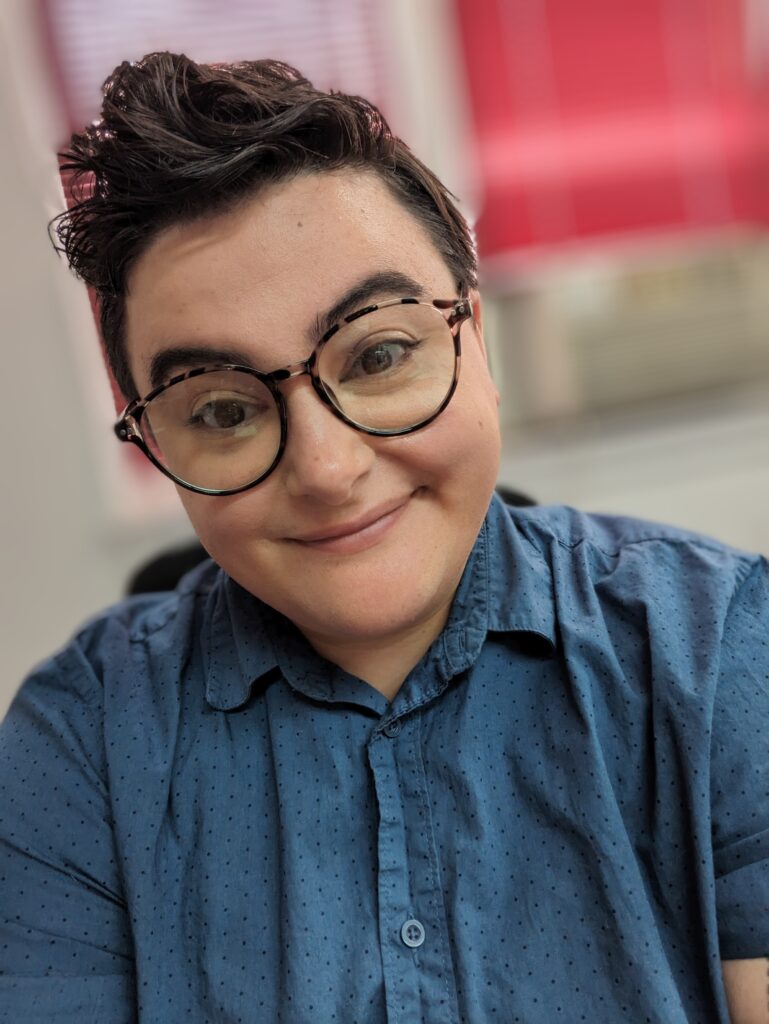 a person with short dark hair wearing glasses and smiling wearing a blue button down shirt and in the background a pink and red display