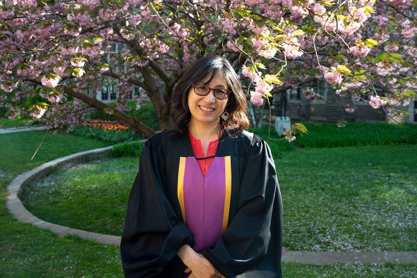 a woman with shoulder length brown hair wearing glasses and smiling, she is also wearing a black robe with a pink and yellow dress underneath, in the background is a blooming magnolia tree