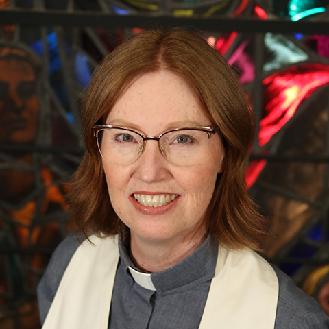 a woman with shoulder length red hair, wearing glasses and smiling, wearing and alb and clergy collar in the background is stained glass in blue and red